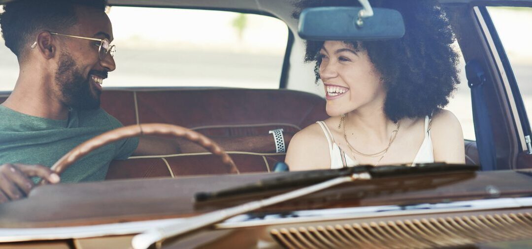 A black man with gold glasses looking to his left at a black woman. They are both smiling and sitting in an old car. 