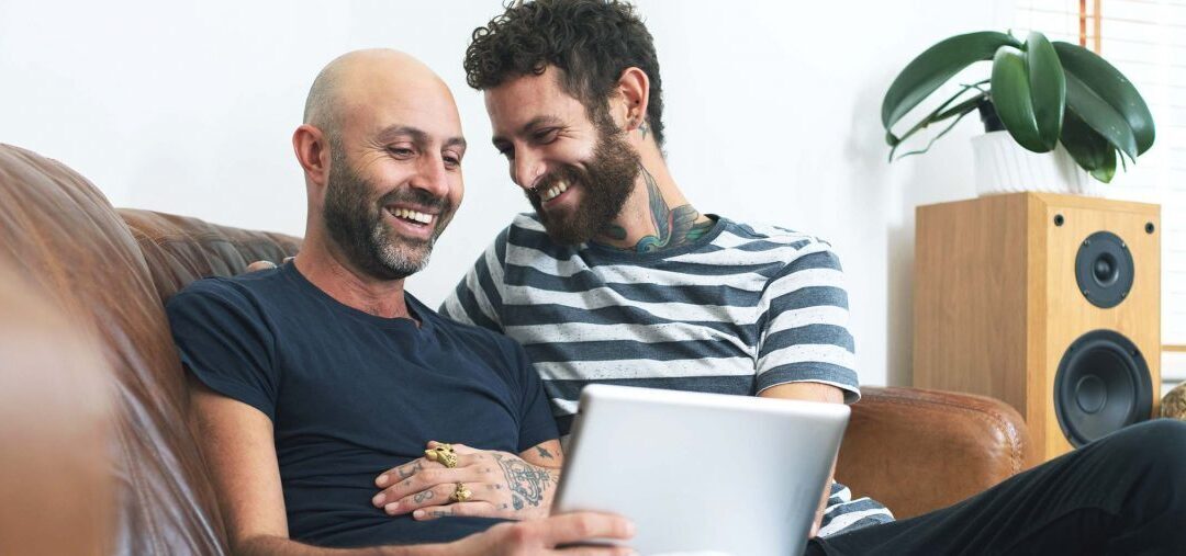 A white bald man with a beard laughing sitting on the couch next to a white man also with a beard. They men are laughing and looking at a laptop. They present as gay together. 