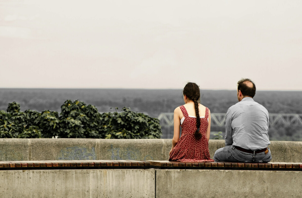 Girl facing out to a lake with a red dress on and a man who is balding sitting next to her. Both of their backs are to the camera and the mood seems down. 