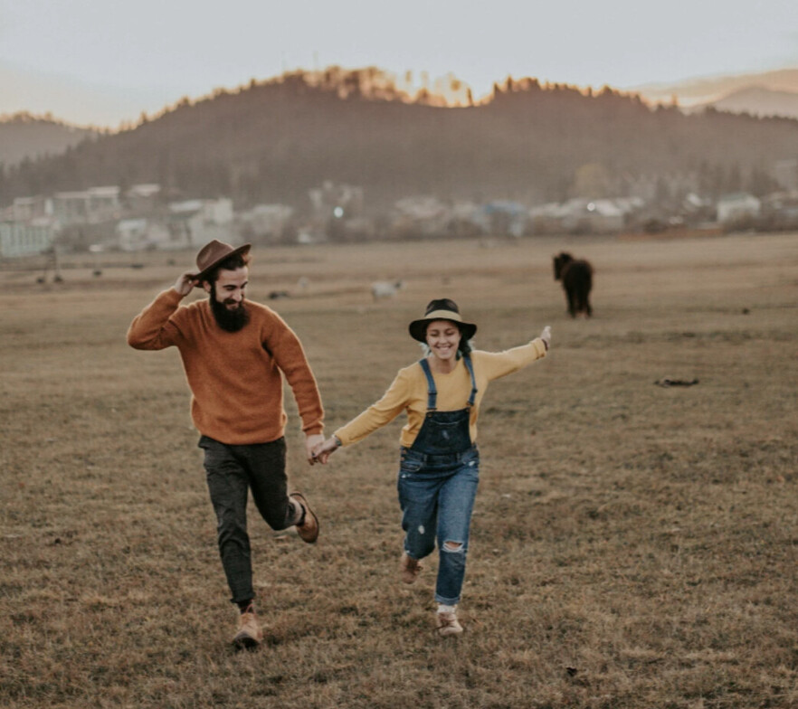 A man with a red shirt running in a field holding his girlfriend's hand. The couple is happy and laughing. 