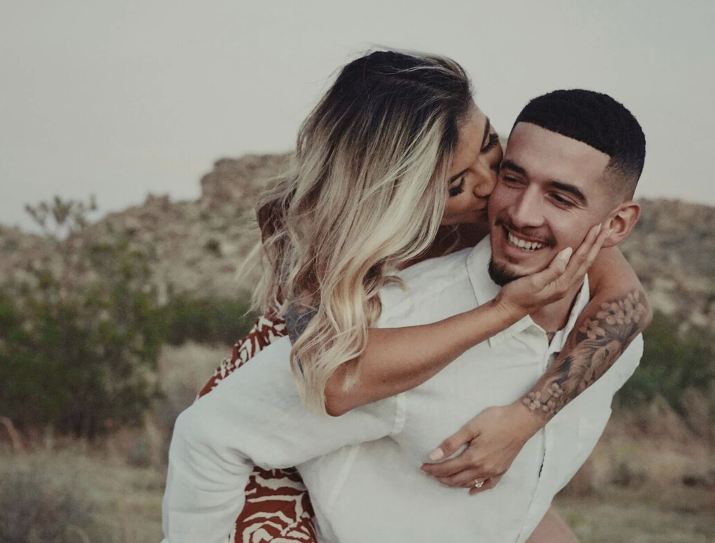 A blonde female kissing her boyfriend. He is giving her a piggy back and has a short hair. The female has an arm tattoo. The couple seems very happy. 
