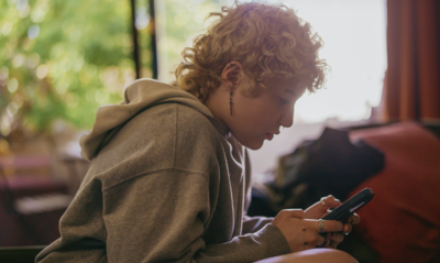 Non-binary presenting female with short blonde curly hair looking at their phone. The tone/vibe is solemn. 