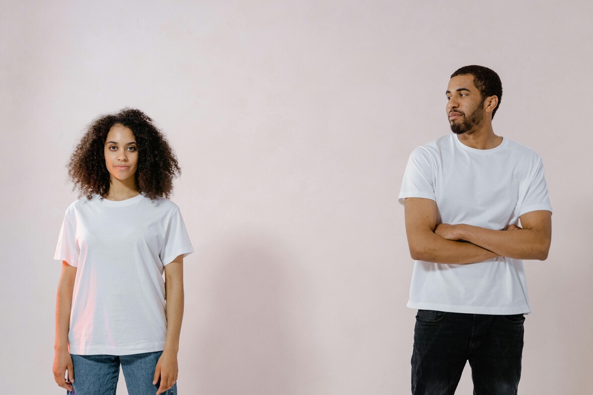 A black woman is standing about 2 feet from a black man. Both people are wearing white shirts and the mood is solemn. The man has his arms crossed.