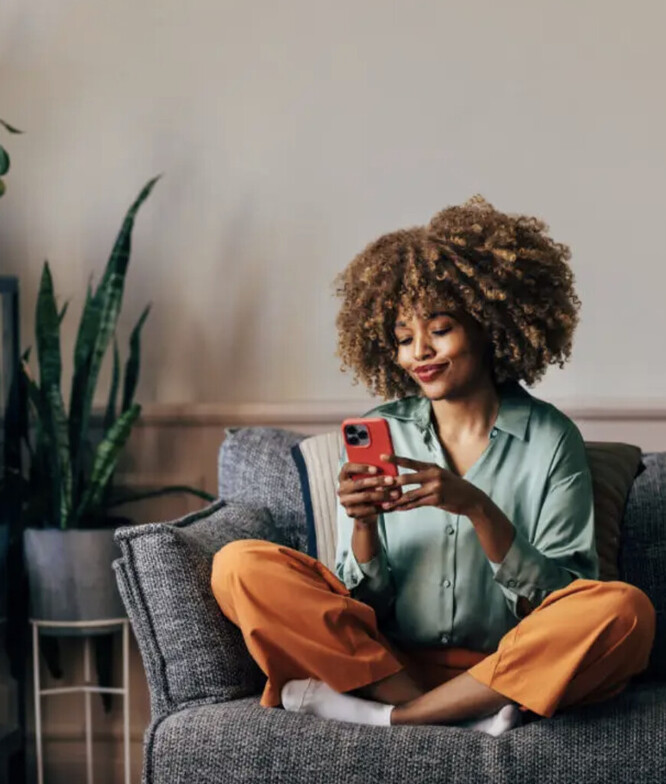 A black woman with afro hair sitting on a couch looking at her phone and smiling. She is wearing a denim shirt and orange pants. 