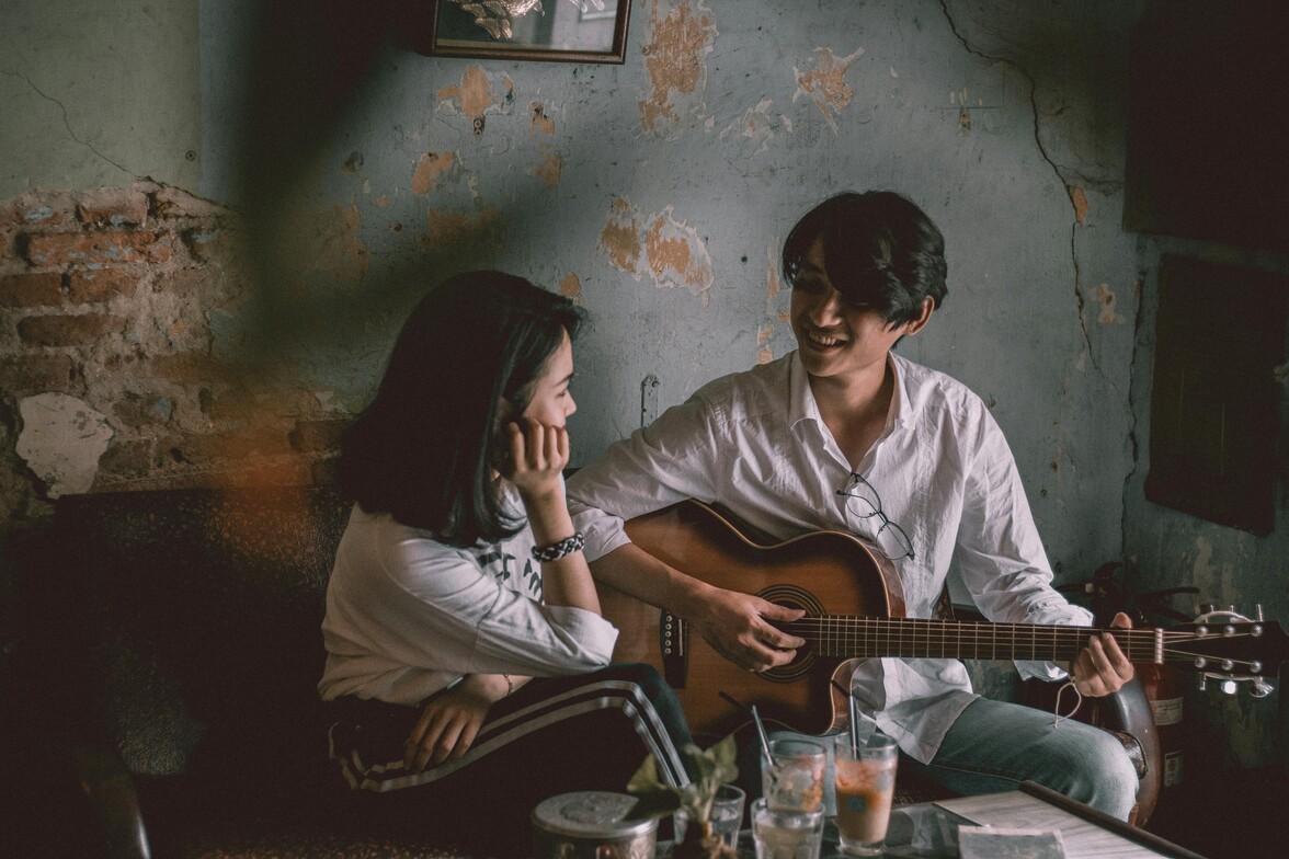 Brown haired women facing an asian man playing a guitar in a living room. 