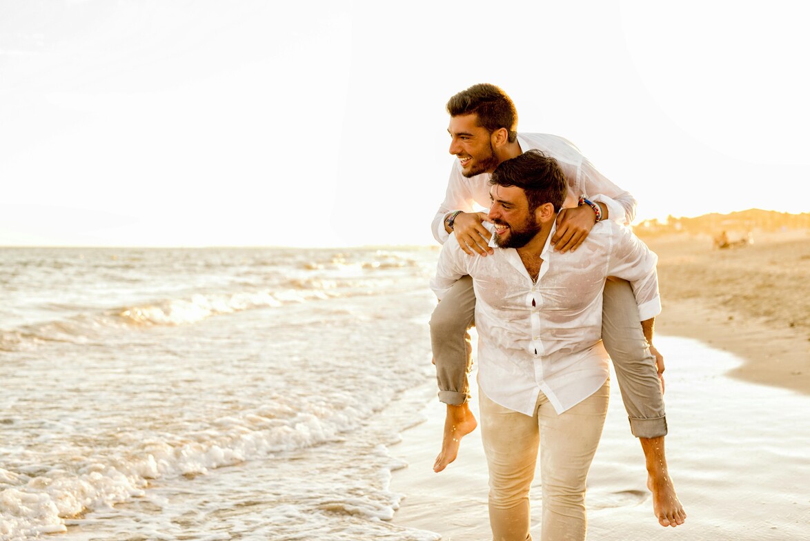 A brown haired man wearing a white shirt on the back of another brown haired man. They are on the beach and both looking to the left. 