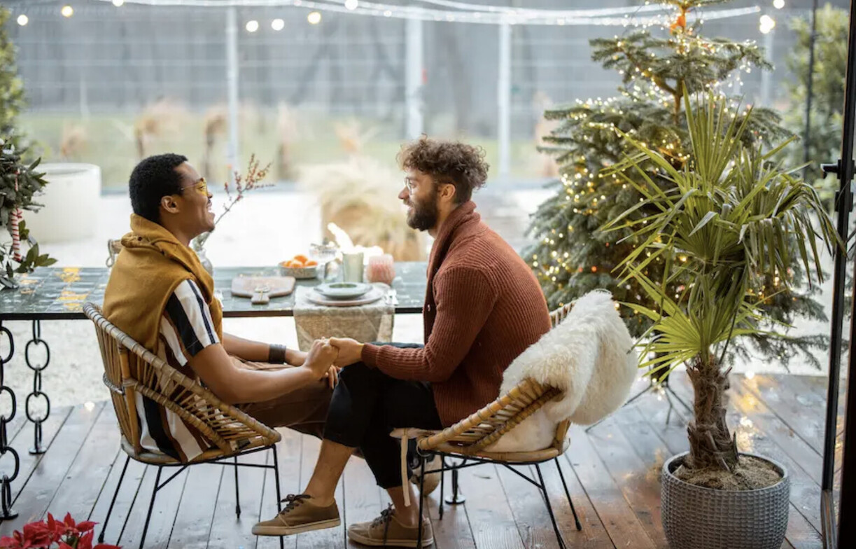 A man with short black hair with a yellow sweater tied over his shoulders holding hands and looking at a man with a burgundy sweater. Both men are looking at each other sitting in a chairs.  