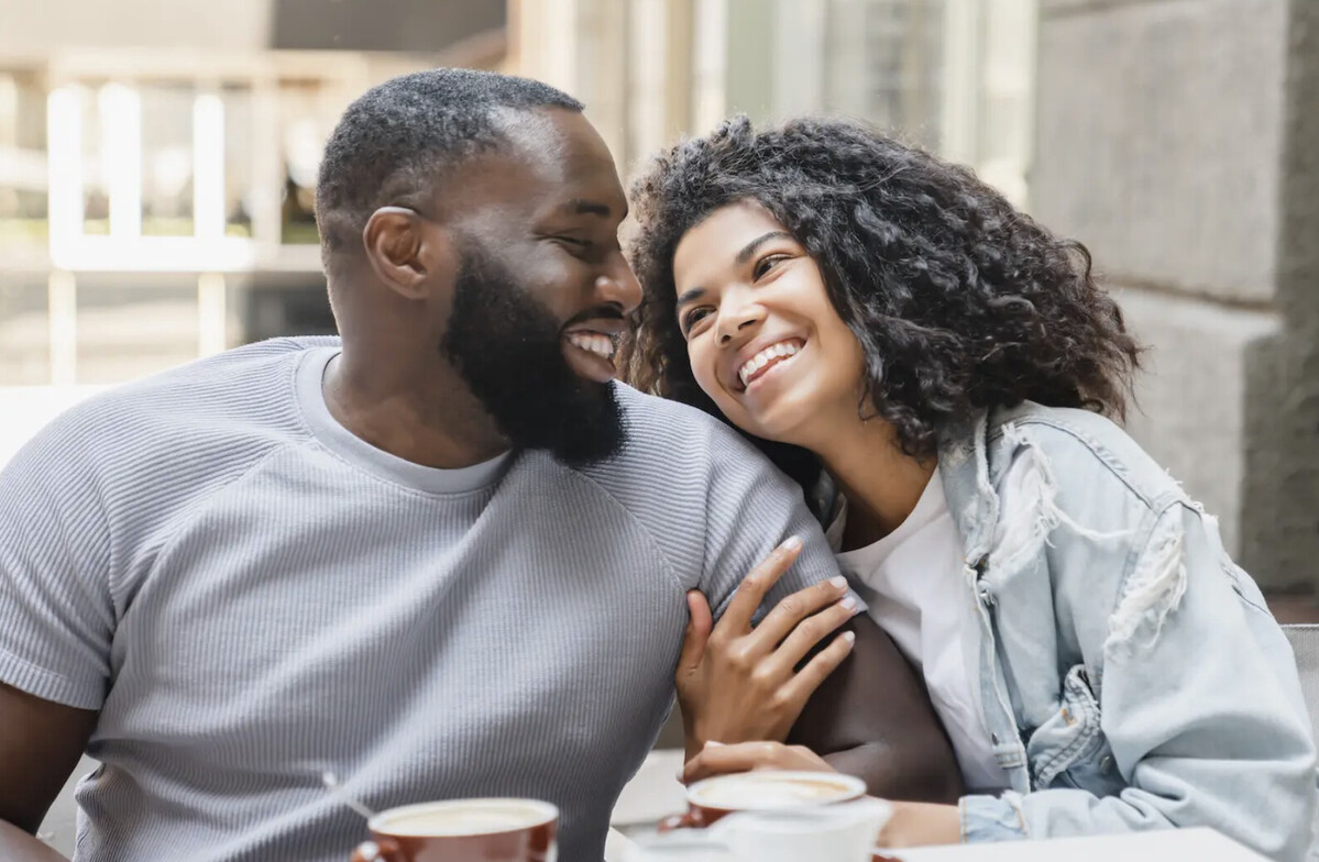 A black man looking to his left lovingly at a black woman holding his arm