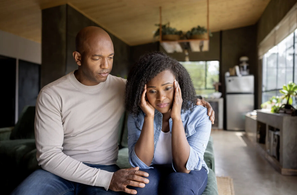 A black man in a white shirt with his arm around a black woman in a blue shirt. They both look sad. 